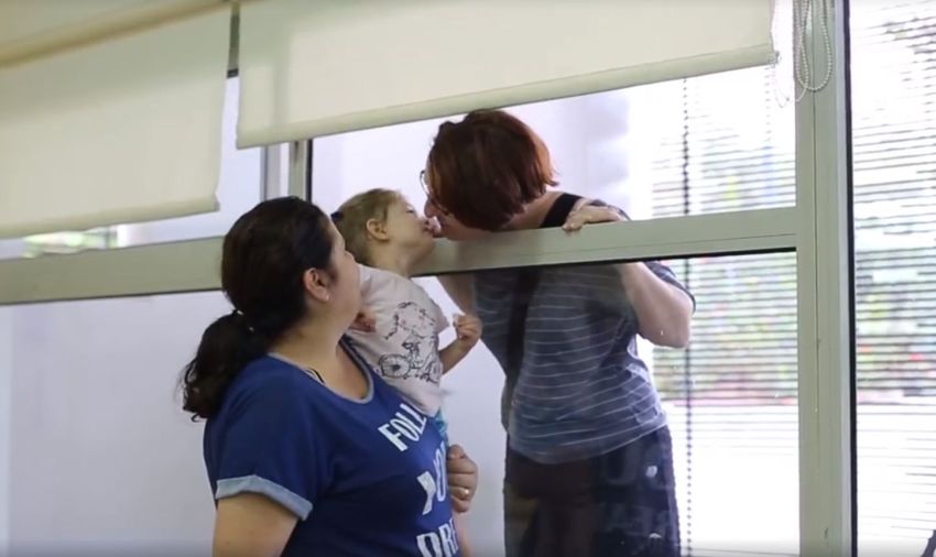 אמא נושקת לבתה דרך החלון, המטפלת מרימה את הילדה שתגיע לחלון
