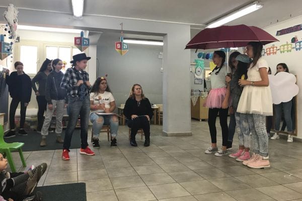 ילדות עומדות מתחת למטריה ומציגות בפני ילדים ומבוגרים
