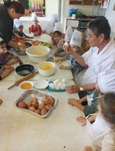 פעוטות בכובעי טבחים יושבים מסביב לשולחן ואישה מבשלת עליו בקערה על השולחן ביצים ועוד פריטי מזון אישה נוספת מגררת גזר על פומפיה