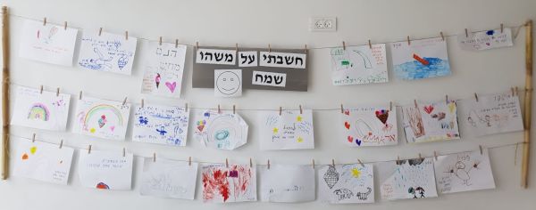 'חבלים' תלויים על קיר ועליהם מוצמדים באטבים ציורי ילדים ומעל הכותרת - חשבתי על משהו שמח