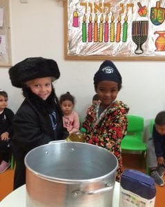 ילד וילדה לבושים כמו חסיד ורבנית ליד סיר גדול מחזיקים בקבוק שמן מעליהם שלט חנוכה שמח
