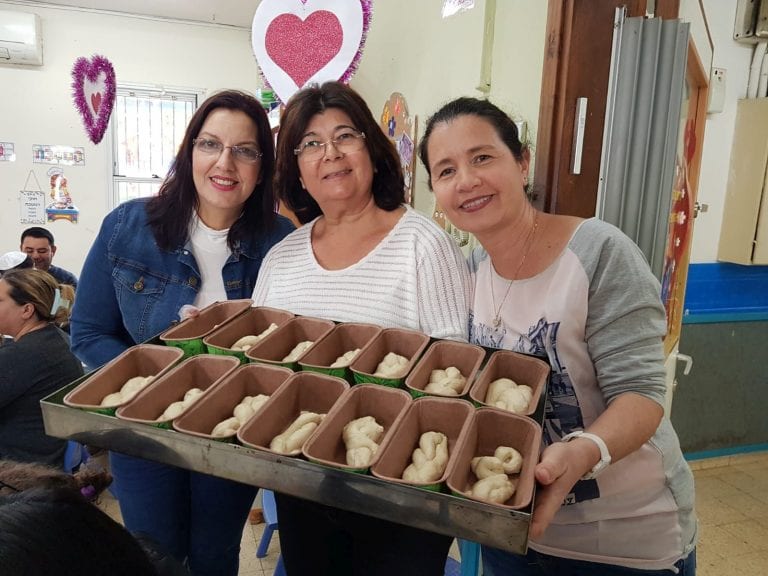 שלוש נשים מחזיקות קרטון ובו תבניות קטנות עם חלות לפני אפיה
