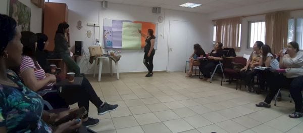 אישה עומדת ליד לוח ונשים יושבות במעגל ומקשיבות