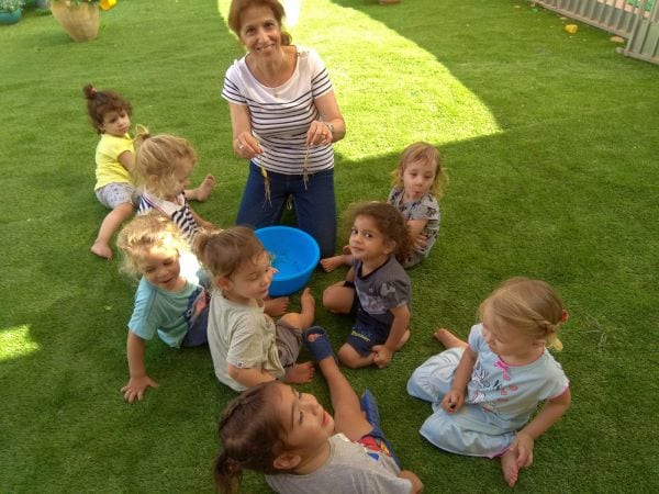 מישל ברבי ופעוטות יושבים על הדשא במעגל ובאמצע קערה כחולה עם הגידולים מהשדה